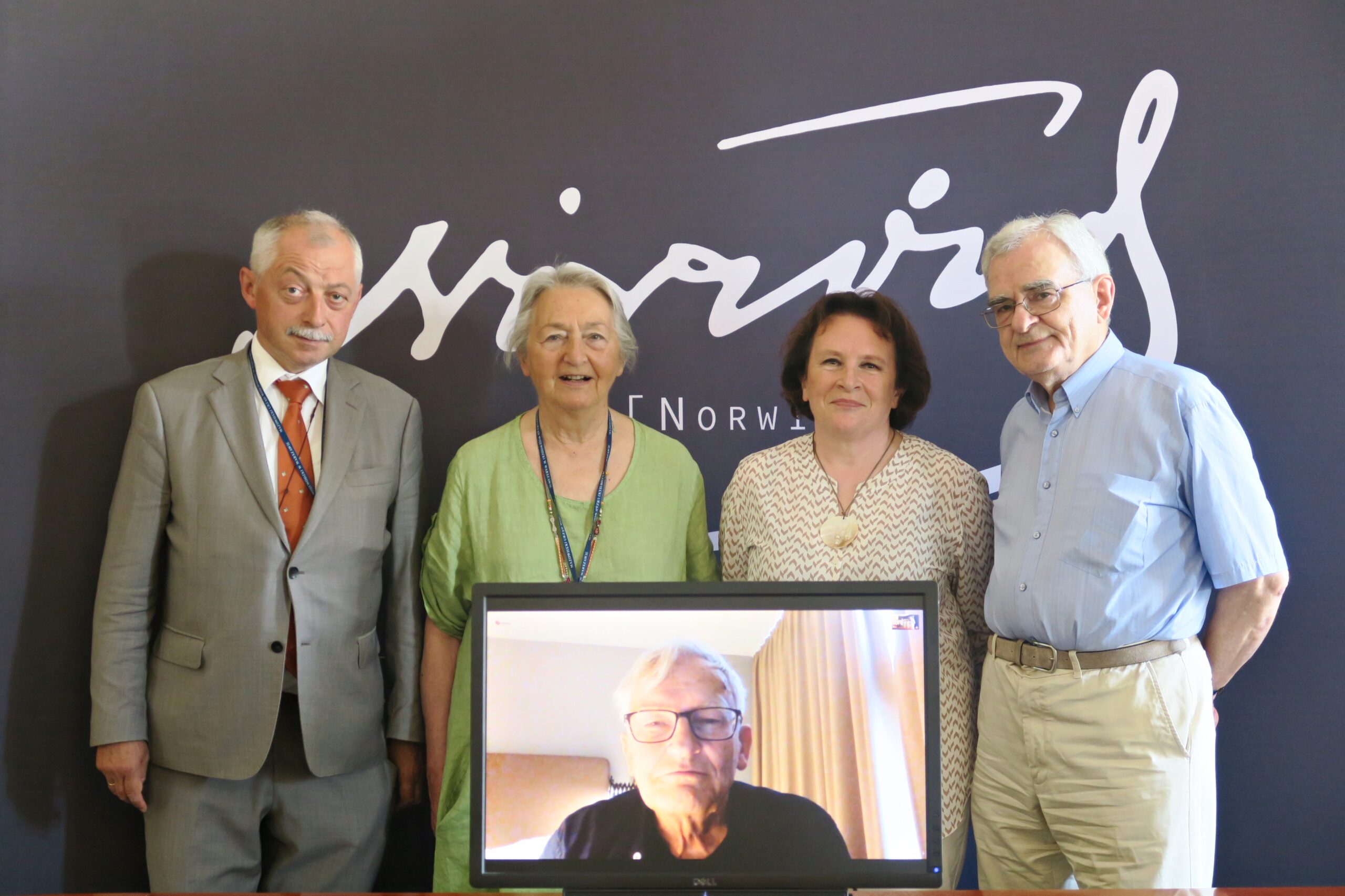 Cztery osoby stoją na tle ścianki z logo Nagrody Norwida. Przed nimi stoi monitor, na którym widać twarz jednej osoby